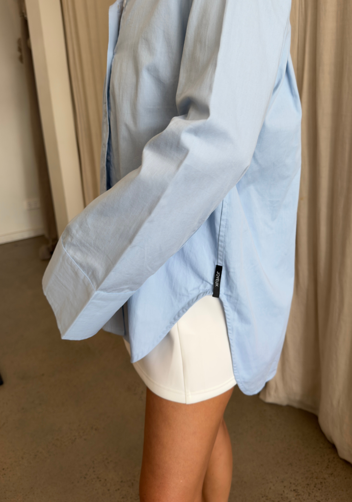 A person wearing an oversized light blue button-up shirt and white shorts, standing indoors in front of beige curtains.