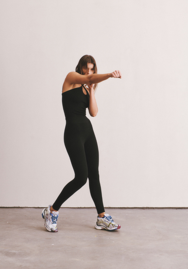 Woman in a black jumpsuit posing against a white wall