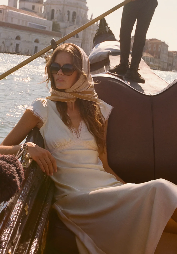 Woman in a white dress and sunglasses sitting in a gondola on a canal with buildings in the background.