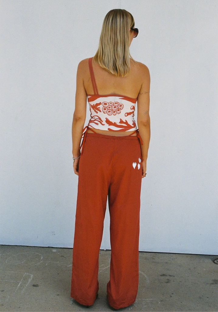 Person wearing a white top with red floral patterns and rust-colored pants against a white wall.